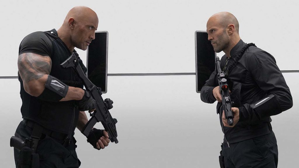 Hobbs and Statham facing one another with guns at the ready and threatening looks. Movie Review