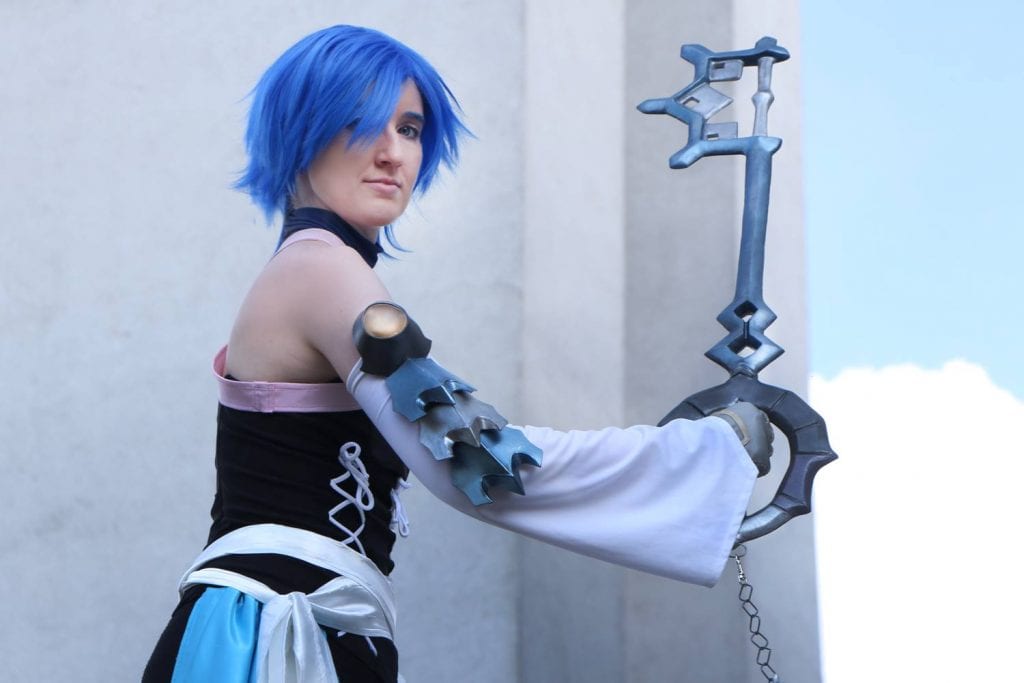 akela dressed as aqua from kingdom hearts birth by sleep. silver keyblade, blue wig, long white sleeves starting from below shoulder with arm guard, black costume with pink white silver and blue accents.