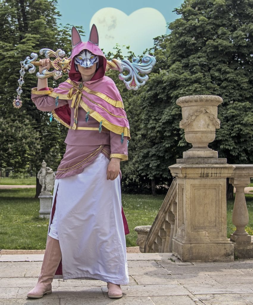 akela dressed as fortune teller Ava from Kingdom Hearts Union X with silver wolf mask, ping and gold top, white skirt, pink boots, large keyblade with garden behind.