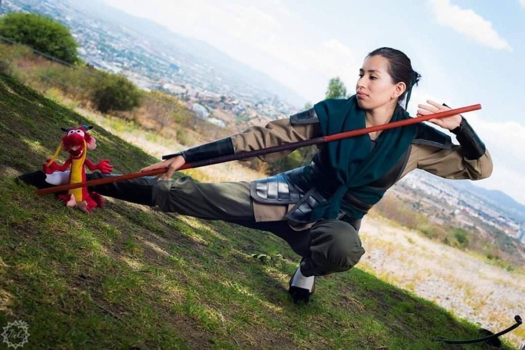 bree dressed as Mulan in her warrior uniform as Ping from disney animated film Mulan. She is holding a staff, Mushu on the ground and cosplayer is in a low pose