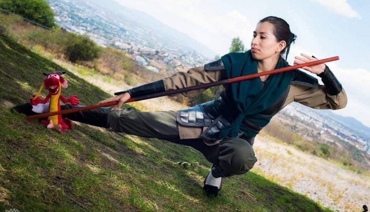 bree dressed as Mulan in her warrior uniform as Ping from disney animated film Mulan. She is holding a staff, Mushu on the ground and cosplayer is in a low pose