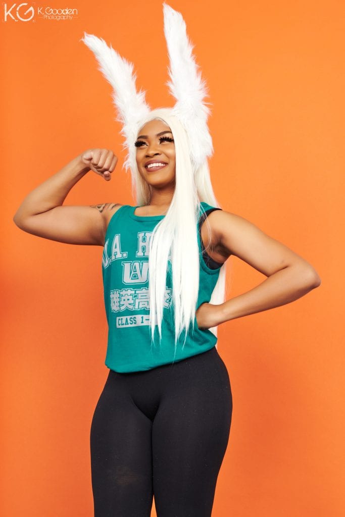 Casual Mirko the Bunny Hero from My Hero Academia. cosplay includes tall white fluffy bunny ears. green MHA tank. black leggings. tall strong pose and huge smile in front of an orange background.