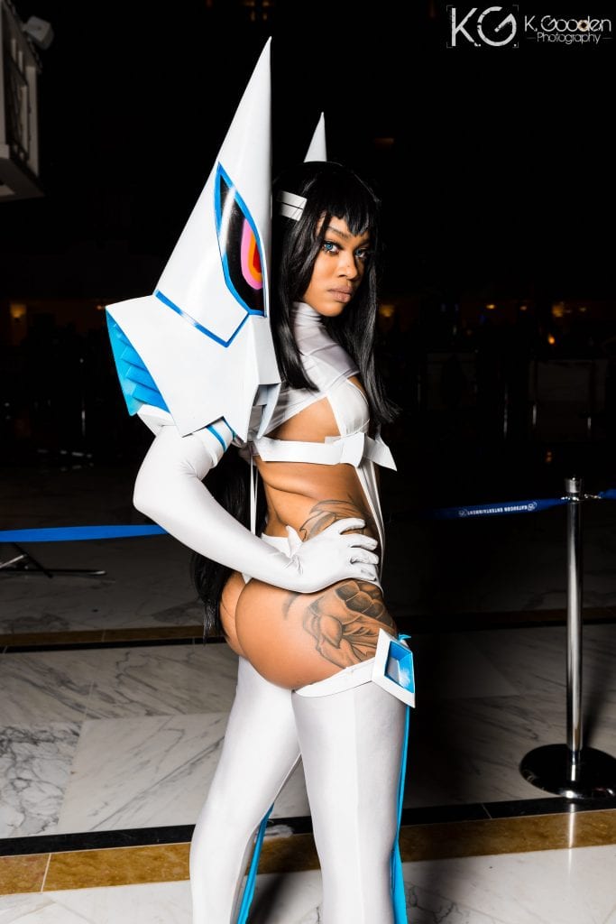 Satsuki Kiryuin from Kill la Kill. Large props off the shoulders that are tall and pointy, white suit thatt covers the front and a little of the back, white stockings, white headband, blue contacts. Cosplayer standing nearly turned to the back looking over her shoulder with hands (long white arm length gloves) on her hips. 