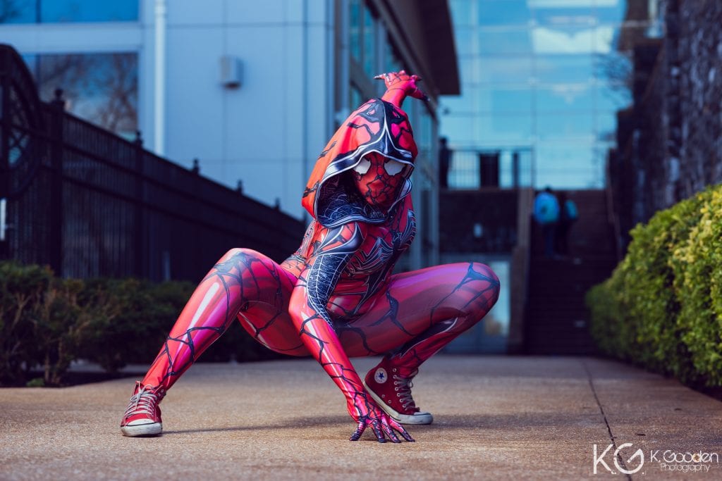 SpiderGwen + Carnage mashup from Spider-Man. cosplay is black and red suit with hood and red sneakers. she's in a deep lunge pose with one hand forward and fingertips on the floor and the other hand is stretched out to the back.
