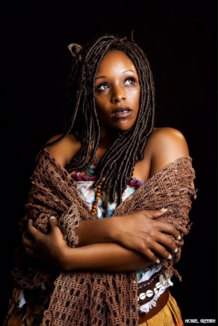 Francheezy dressed as Tia Dalma from Pirates of the Caribbean with a brown shawl, dark brown tight dreads, yellow skirt, maroon trimmed corset. Her arms are crossed with the shawl.