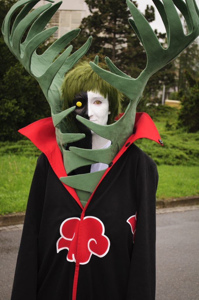 akela dressed as zetsu from naturo shippuuden with akatsuki coat that is black with red clouds embroidered with white borders and red interior. green hair, half white half black face with red and yellow contacts and large green prop protruding from the coat about 2-3 ft tall.